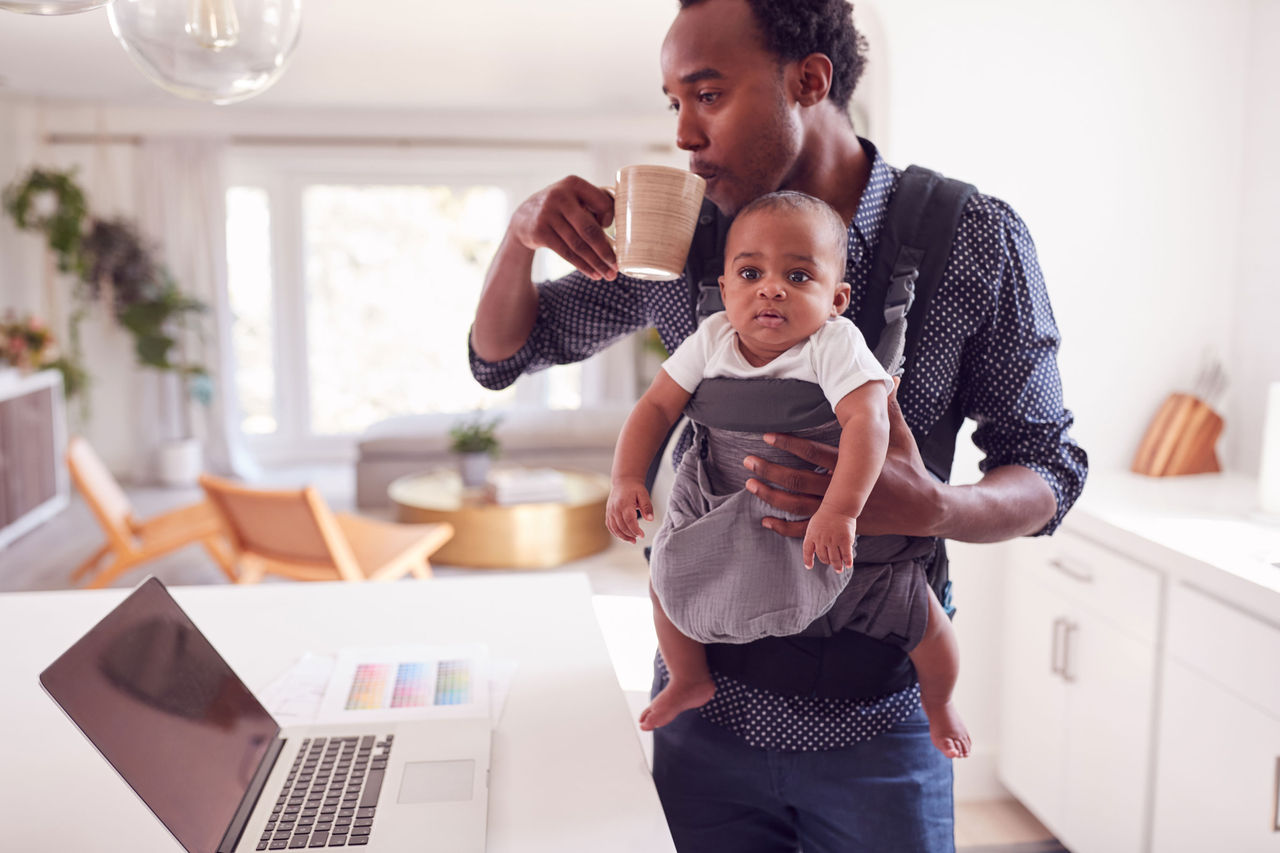 Dad holding a baby in the kitchen while looking at a laptop and drinking from a mug