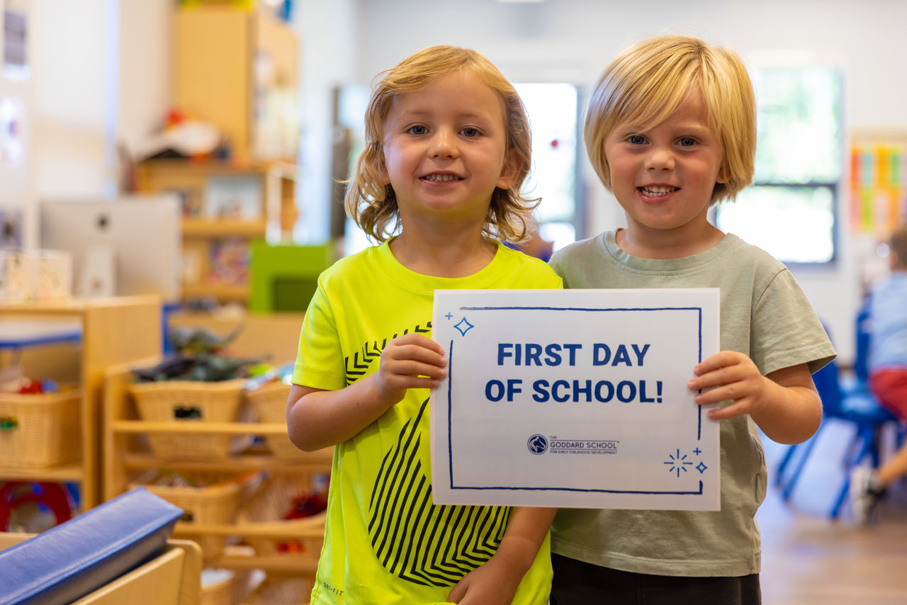 Two smiling preschool children holding a "First Day of School!" sign in a classroom