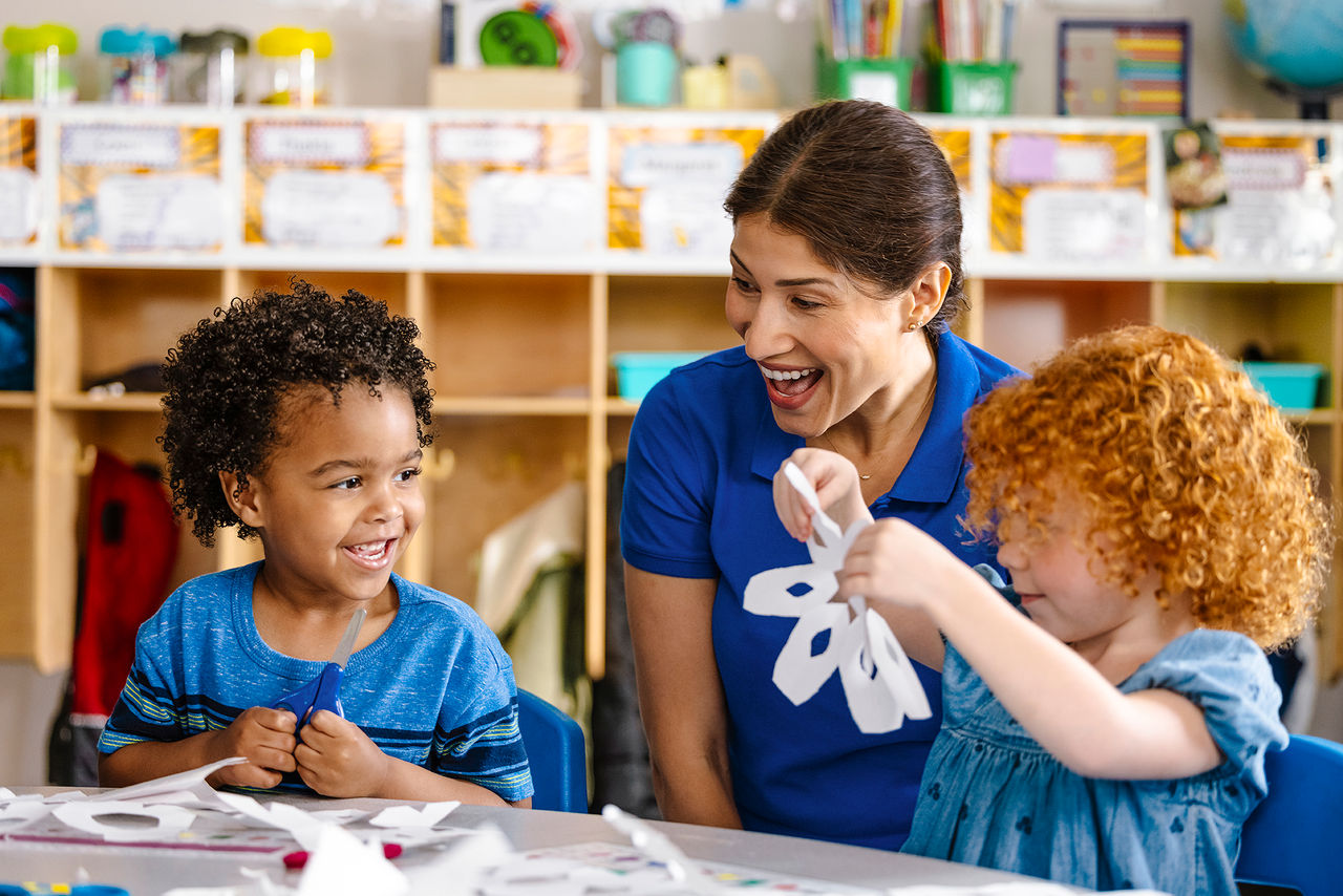 Two preschool children creating paper snowflakes with a teacher.