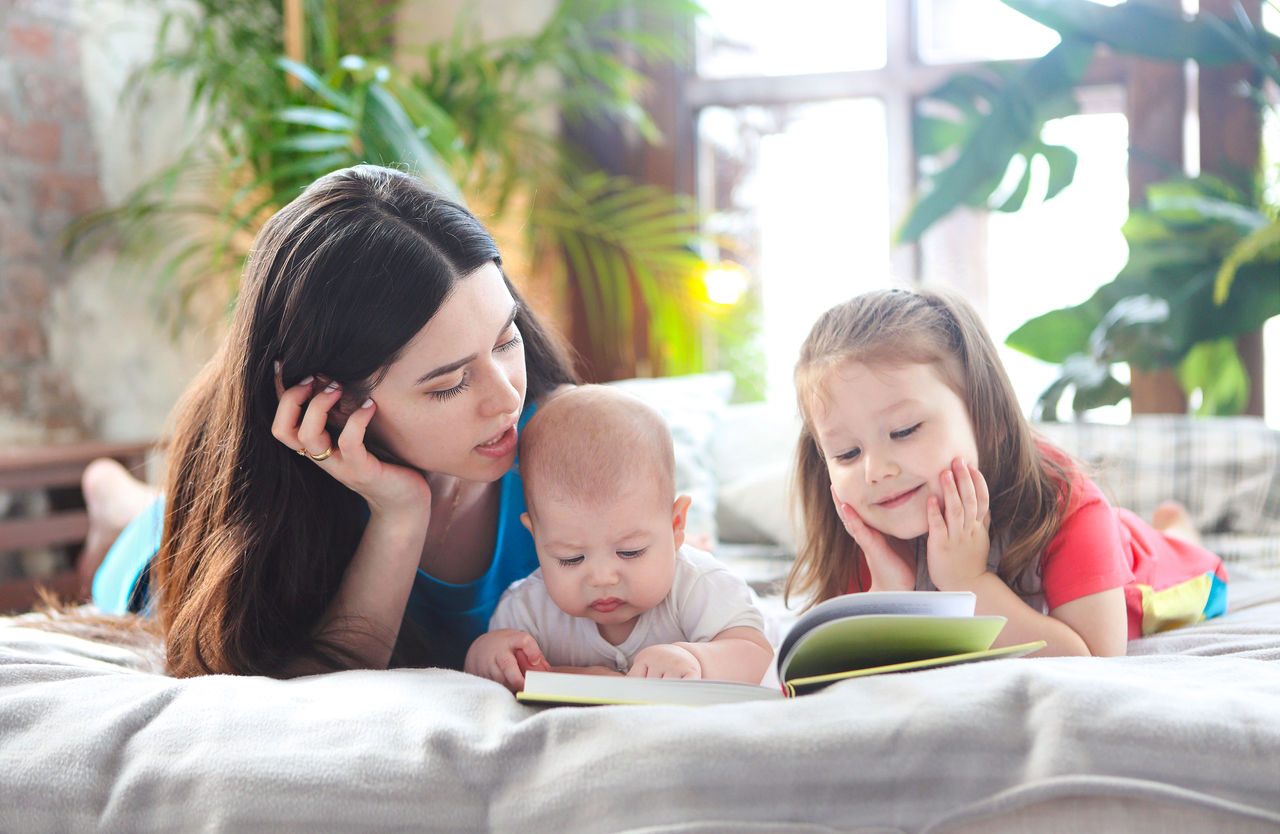 A mom lying on a bed reading a book with her infant and young child