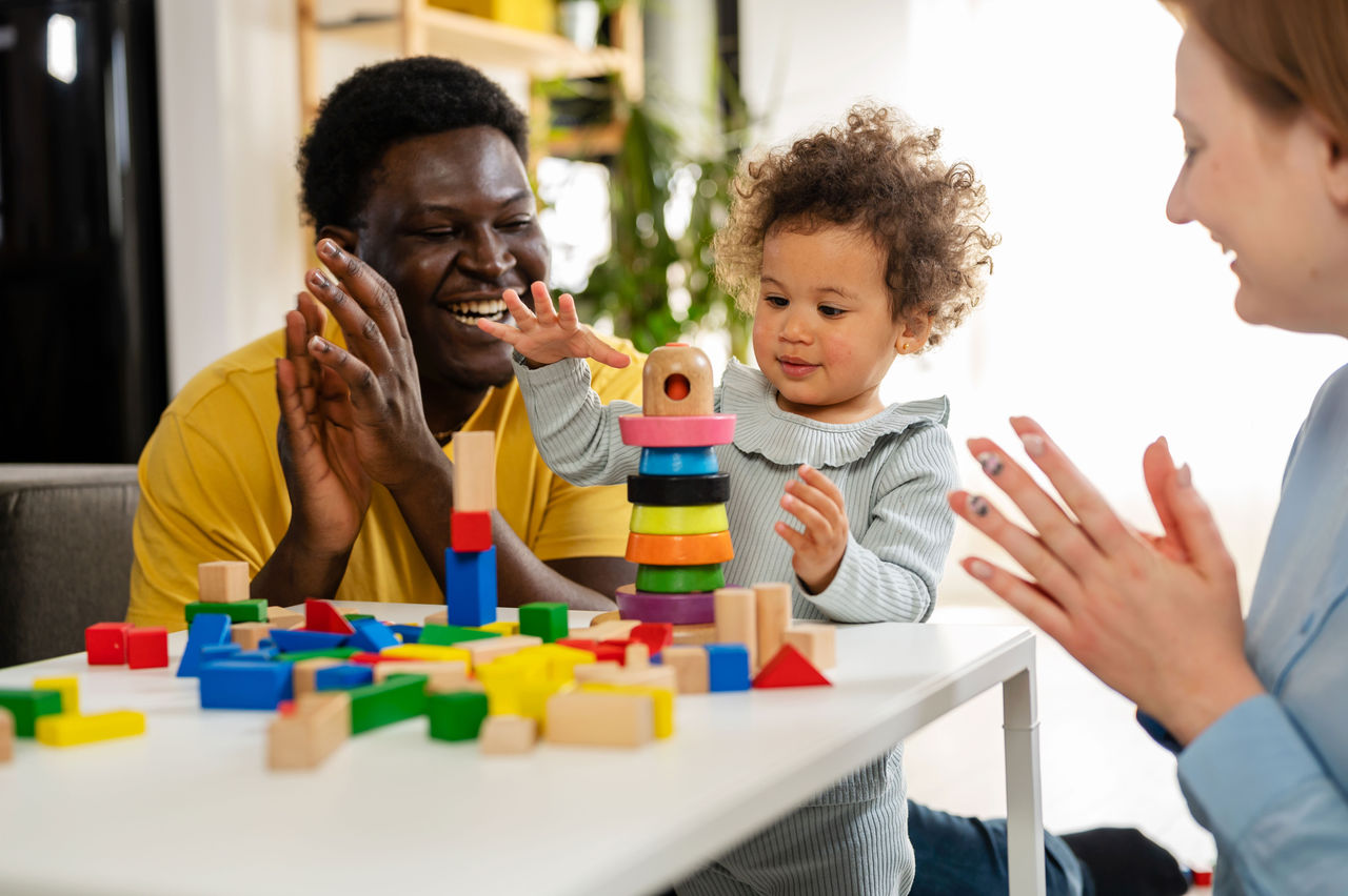 Parents cheering on their baby who is stacking blocks at a table in their home