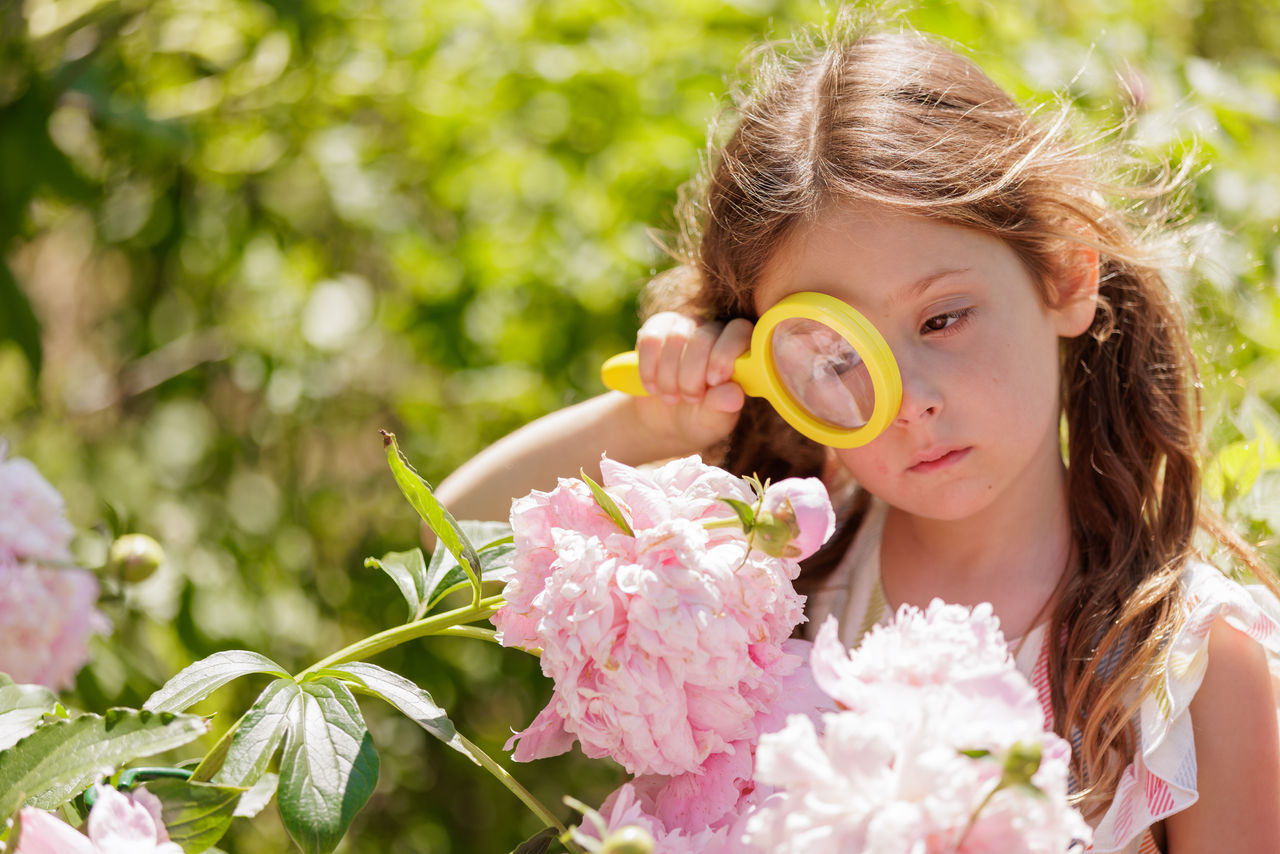 A young girl outdoors looking at a pink flower through a yellow magnifying glass