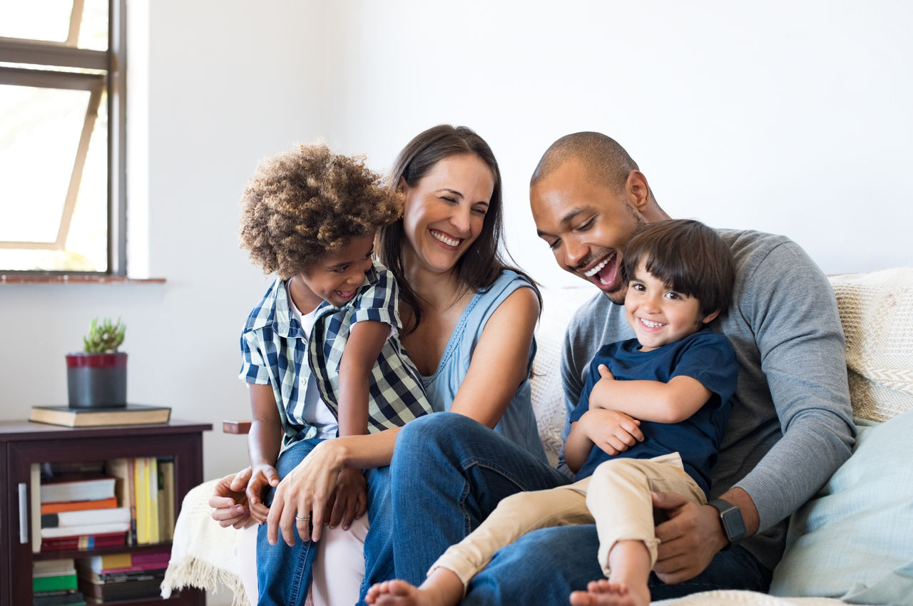 Happy multiethnic family sitting on sofa laughing together. Cheerful parents playing with their sons at home. Black father tickles his little boy while the mother and the brother smile.