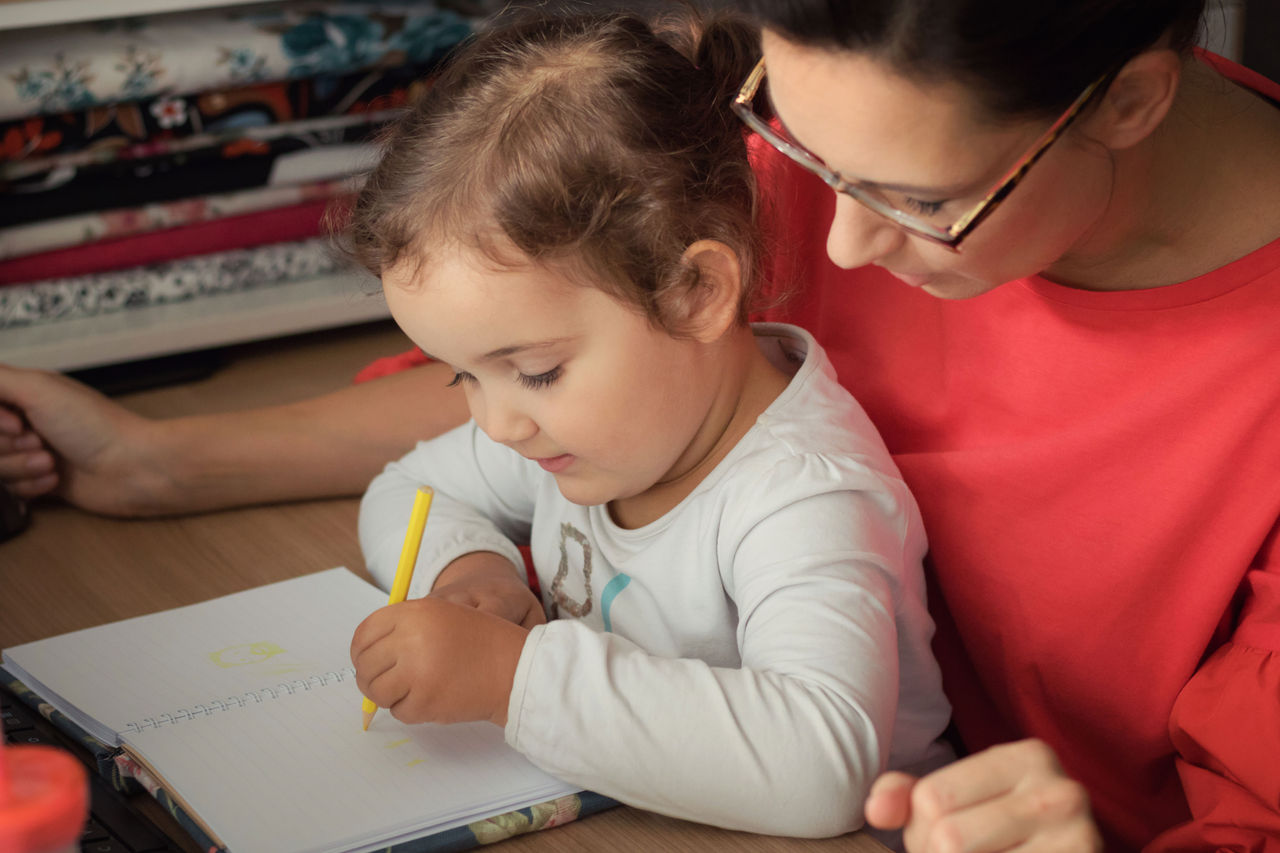 A young child sitting in her mother's lap writing in a notebook