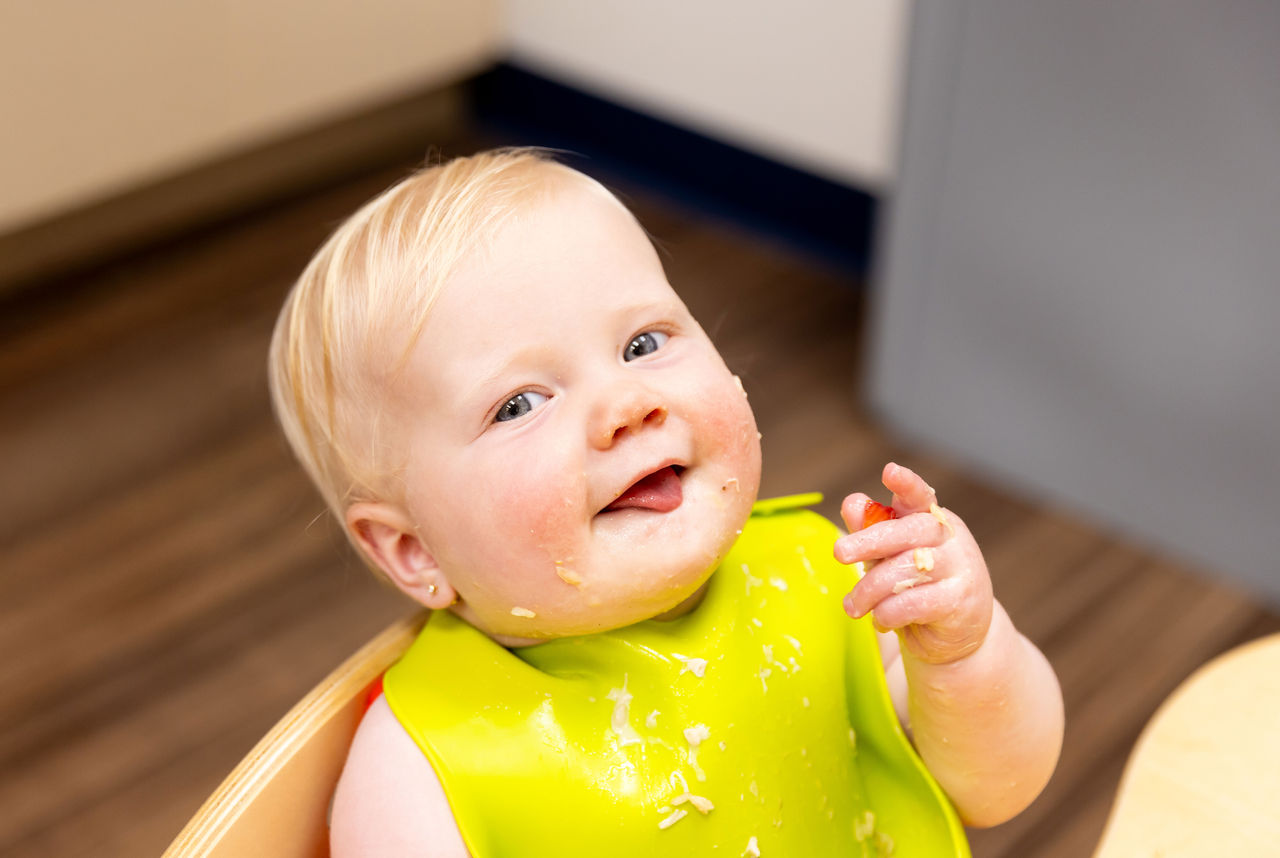 A cute smiling baby wearing a bright green bib with food on his hand, face and bib