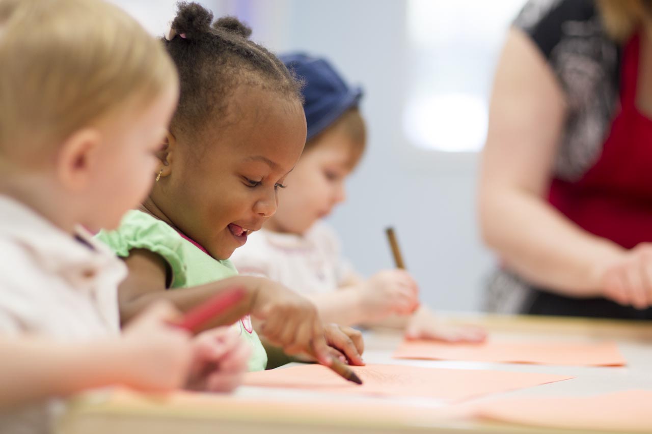 A young child, in a group with others, smiling and holding a brown crayon to an orange piece of construction paper.