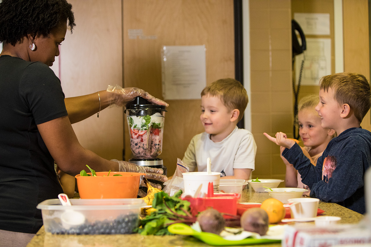 A group of young children watch as a teacher operates a blender with fruit inside.