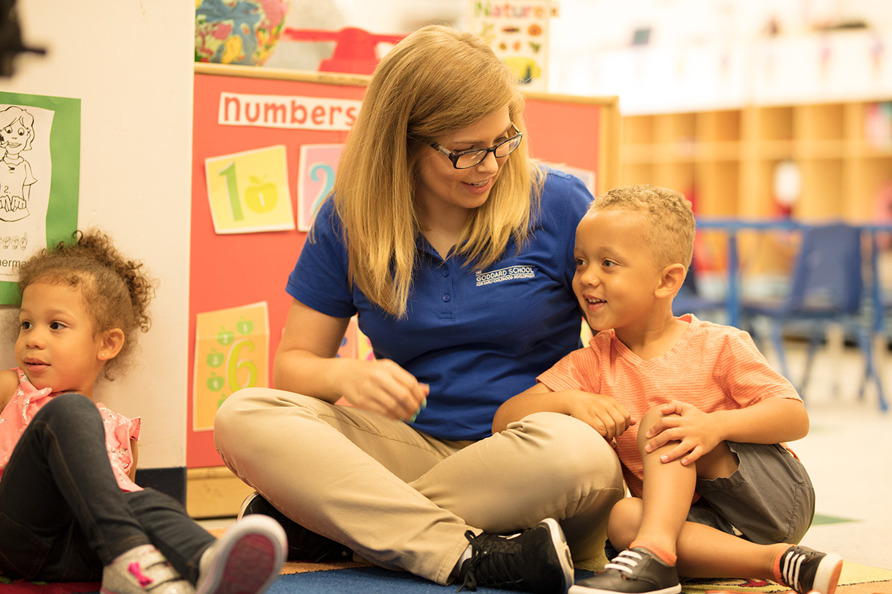 A teacher sitting with her legs crossed on the floor with a young child sitting next to her, smiling and leaning on her.