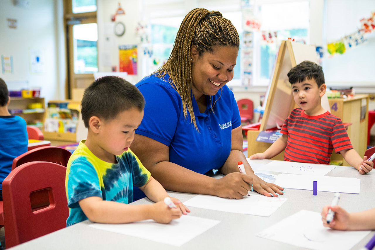 A Goddard School teacher drawing at a table with two young students