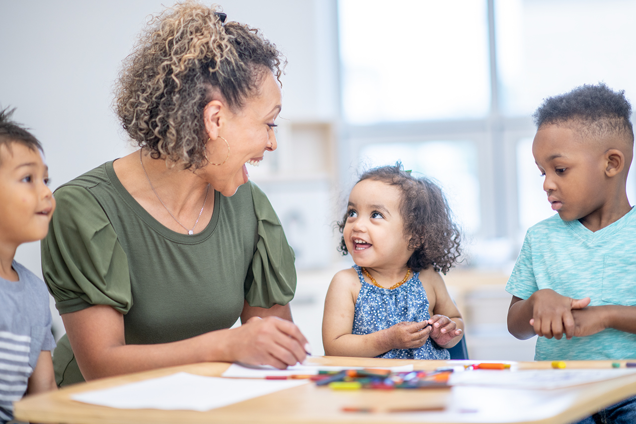 A teacher laughing with a small group of students who are coloring on paper at a table.